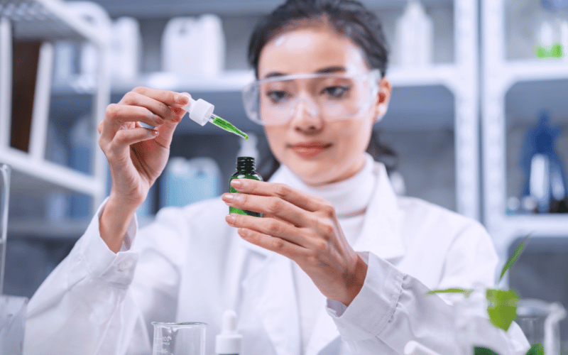 A female researcher in a lab coat examining a green liquid sample in a test tube.